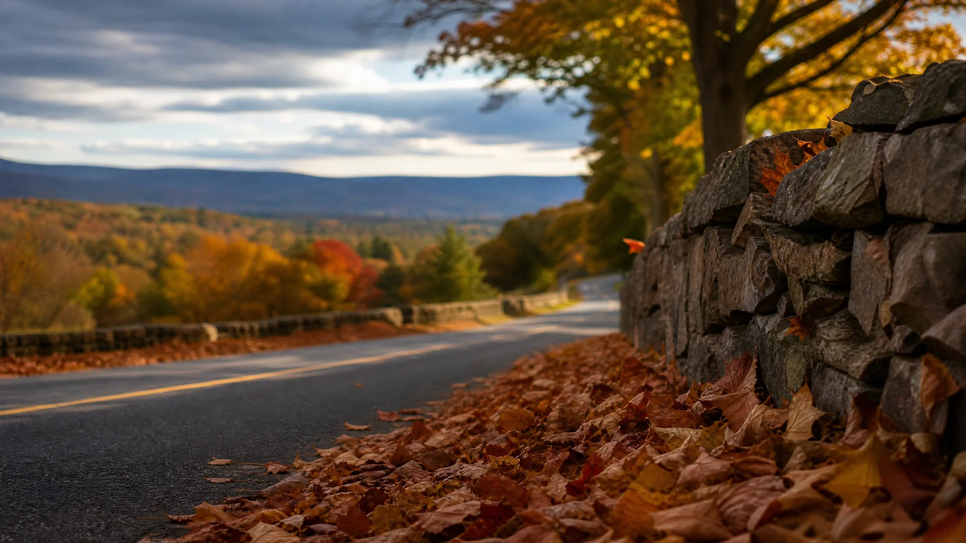 A curving paved road with a yellow center line runs beside a dry-stacked stone wall and a bank of fallen autumn leaves, with distant hills and trees in fall color under a cloudy sky.